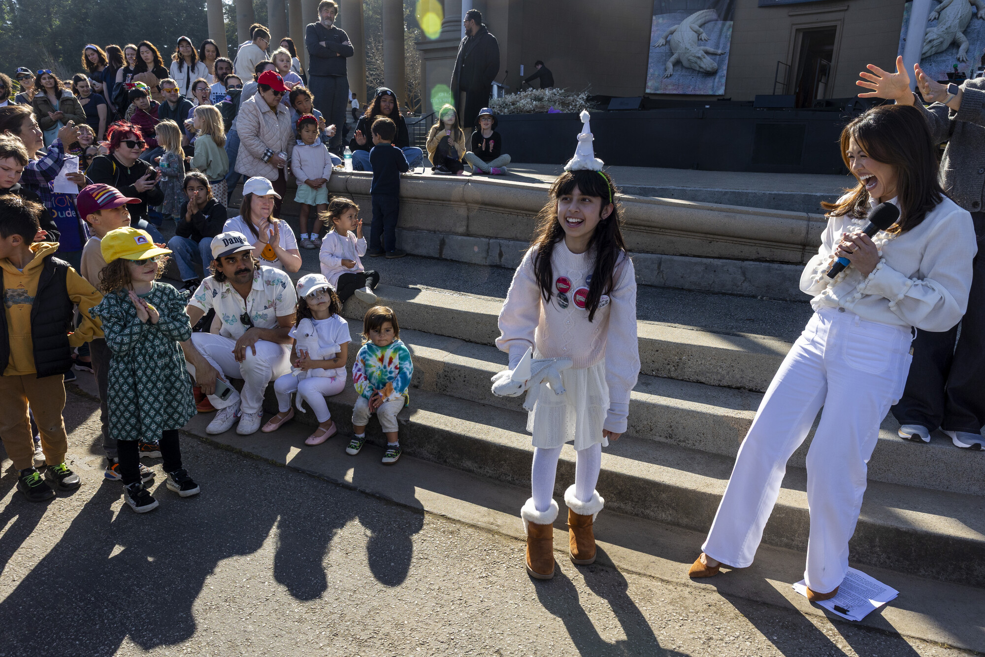 A little girl wearing all white stands smiling, holding a stuffed white alligator as an Asian woman with a microphone smiles adoringly next to her. Children watch on from the sidelines.