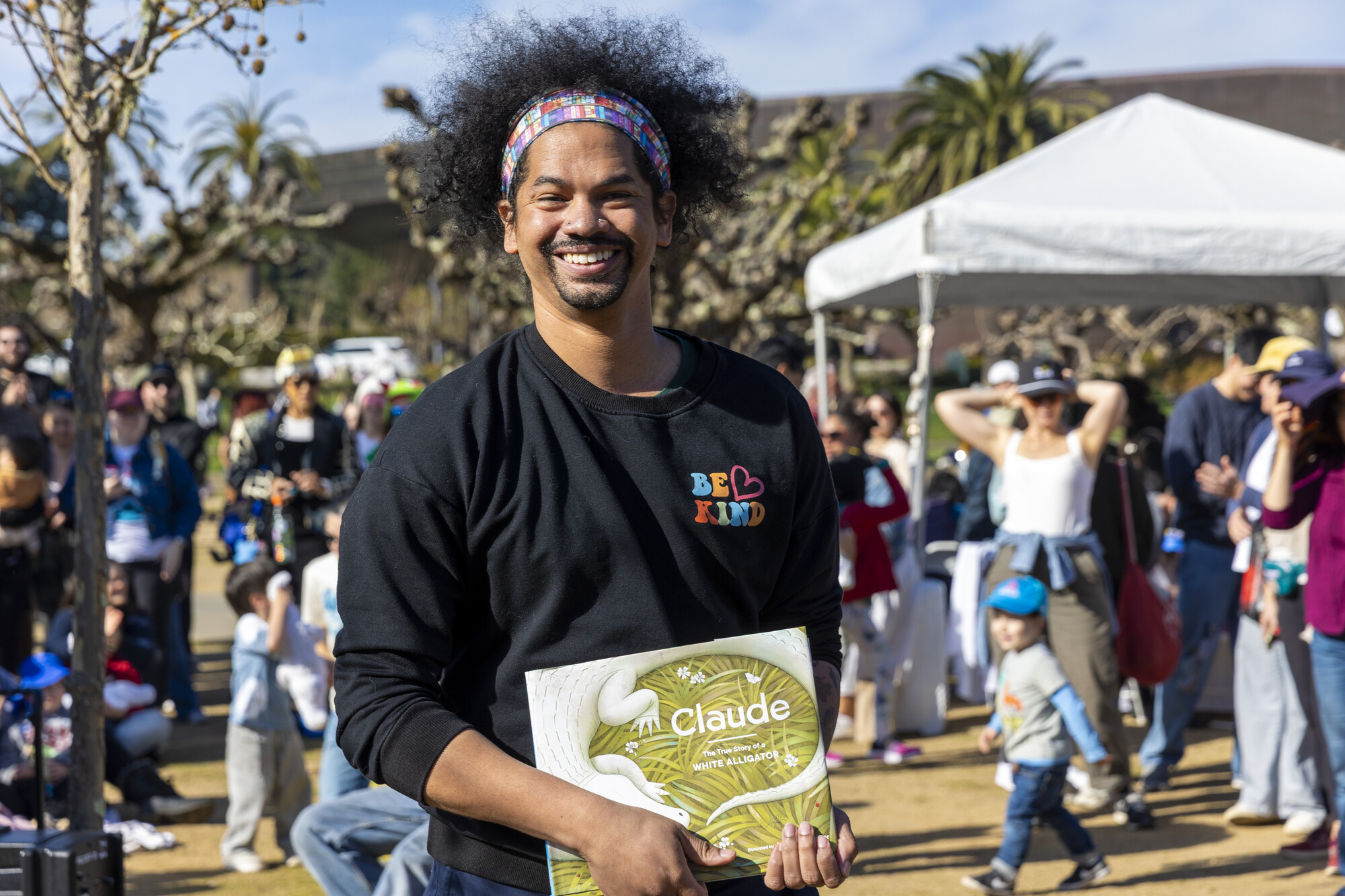 A young Black man with frizzy hair pushed back in a headband smiles and holds a children's book with an alligator on the front. He is standing in a sunny park.