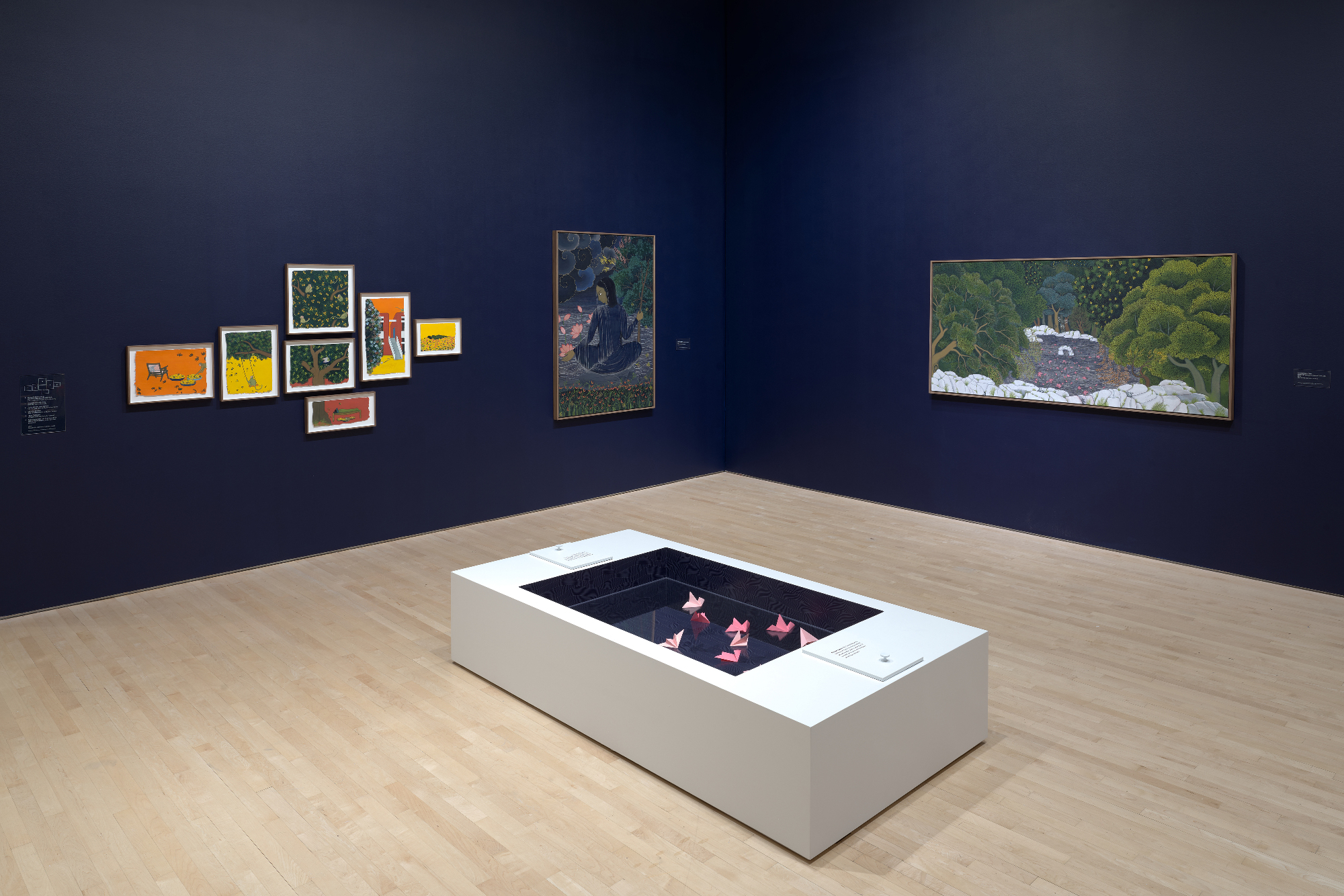 navy walls with paintings and framed works on paper, pedestal with origami in center of room