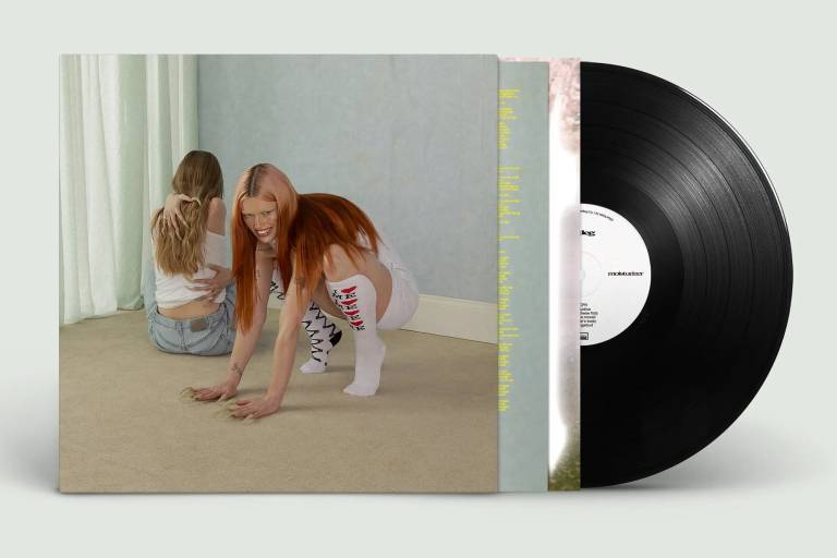 A woman with long reddish hair and long socks crouches in an insect-like stance in a bare living room, while another woman sits with her back turned
