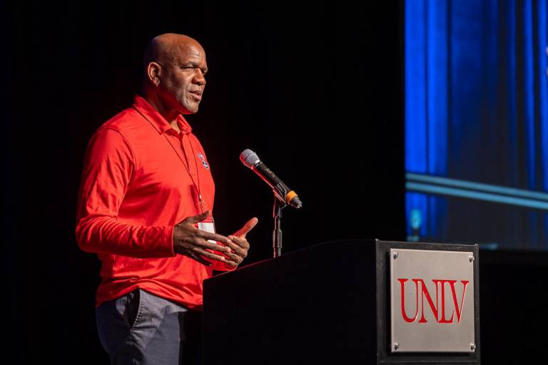 A bald African American man in a red shirt standing at a podium that reads "UNLV."