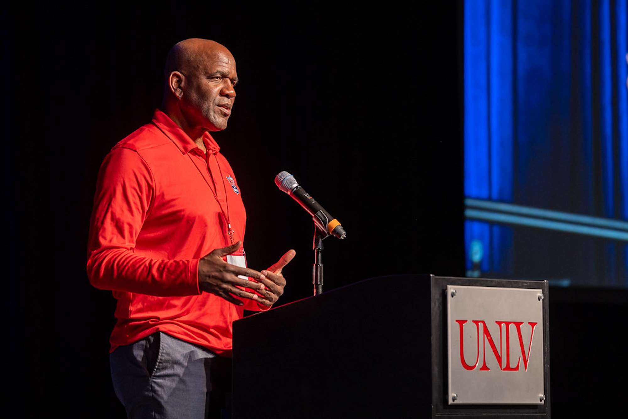 A bald African American man in a red shirt standing at a podium that reads "UNLV."