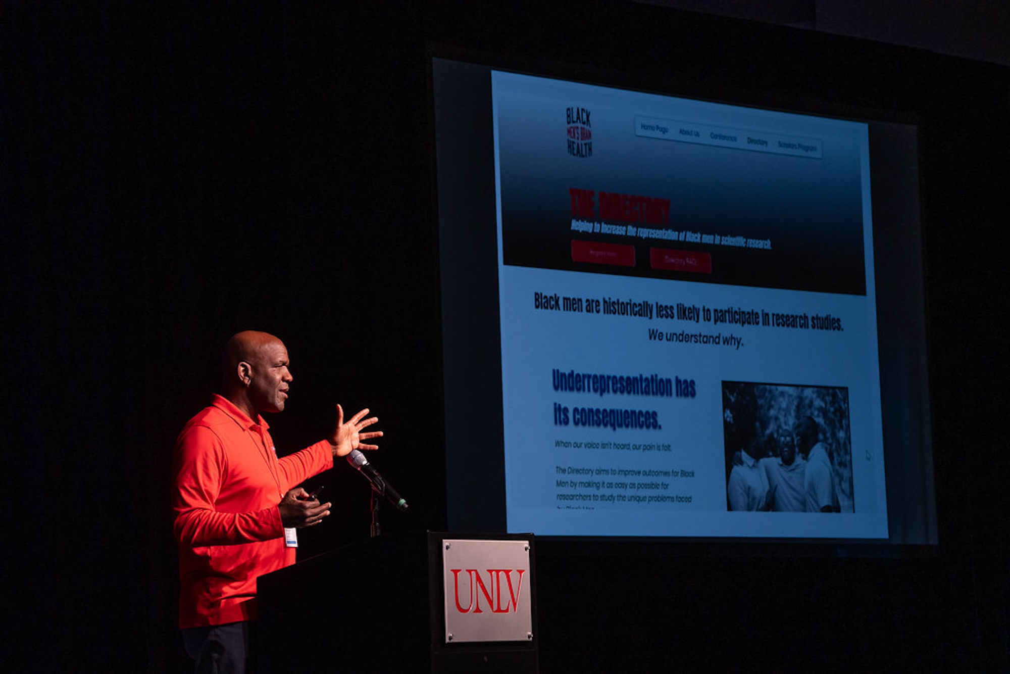 A bald African American man in a red shirt stands on a stage giving a presentation on health outcomes for Black men in America.