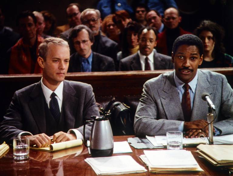 Tom Hanks and Denzel Washington sit behind a courtroom desk strewn with papers and notebooks in a scene from Philadelphia.