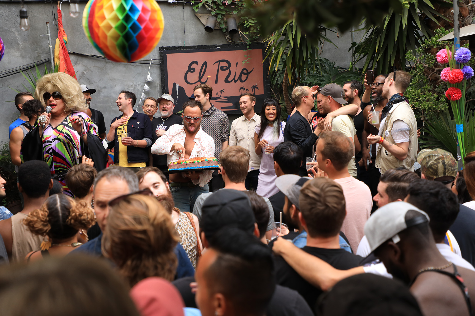 Crowd mingling on a bar's outdoor patio. A pink sign in the back reads, "El Rio."