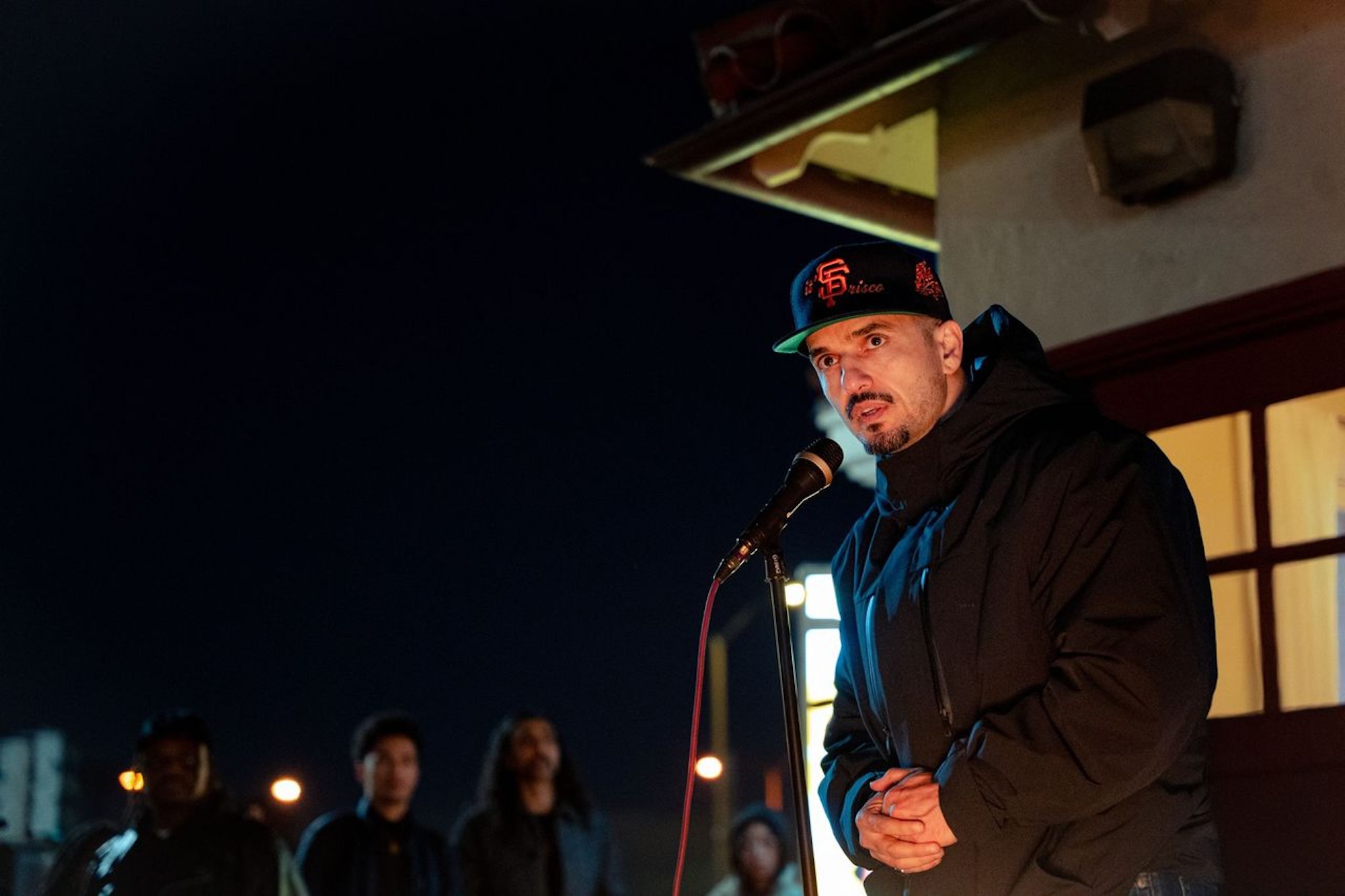 A candid photo of a man with a black and orange SF Giants hat and a black hoodie, as he stands behind a microphone addressing an audience. 