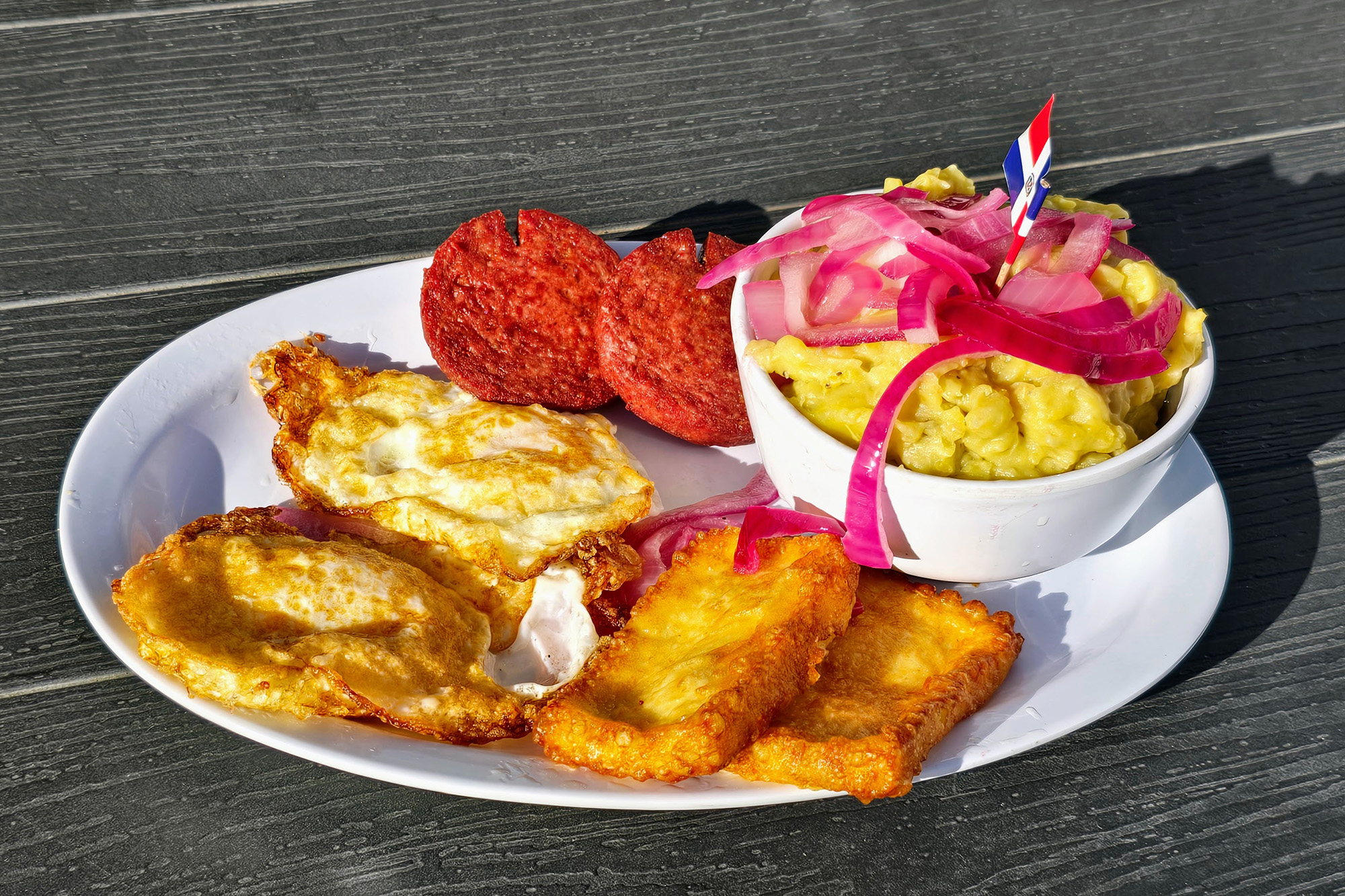 A plate with mashed plantains, fried eggs, and sliced of fried salami.