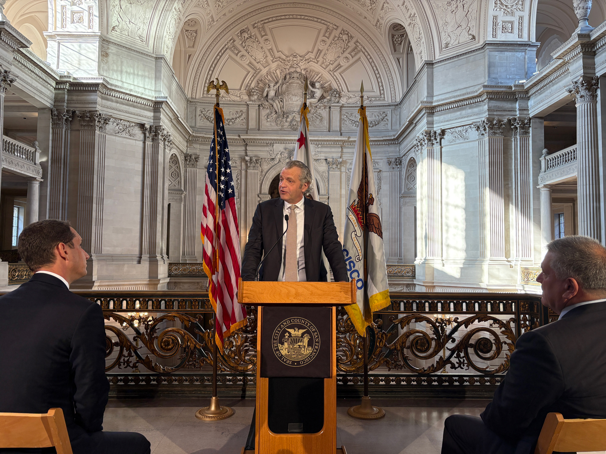 two men seated before standing man at podium in grand rotunda