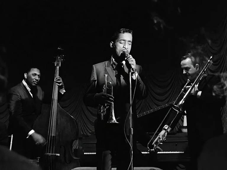 A slendar Black man sings into a microphone, flanked by a bassist and trombonist, while holding a cornet in a black and white image.