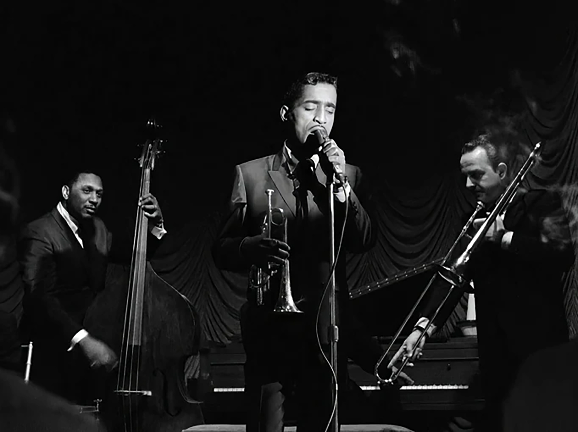 A slendar Black man sings into a microphone, flanked by a bassist and trombonist, while holding a cornet in a black and white image.