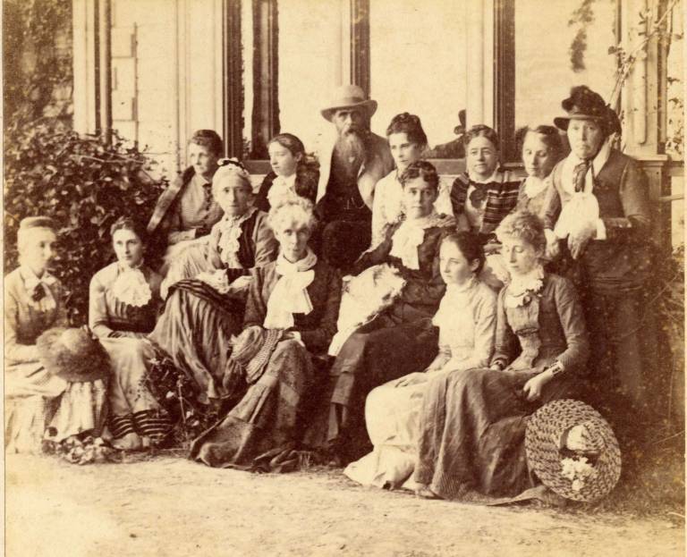 A man is surrounded by women, all in modest 19th century clothing, in sepia toned photo from the 1800s