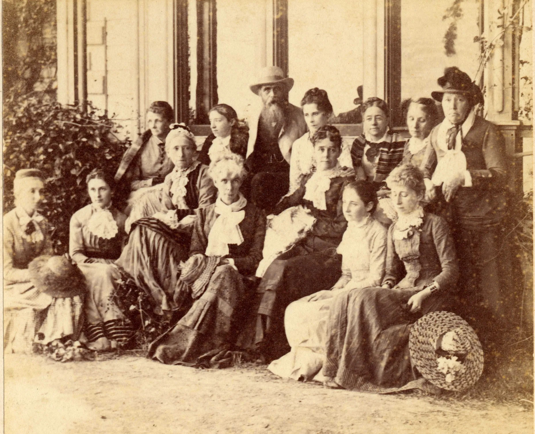 A man is surrounded by women, all in modest 19th century clothing, in sepia toned photo from the 1800s