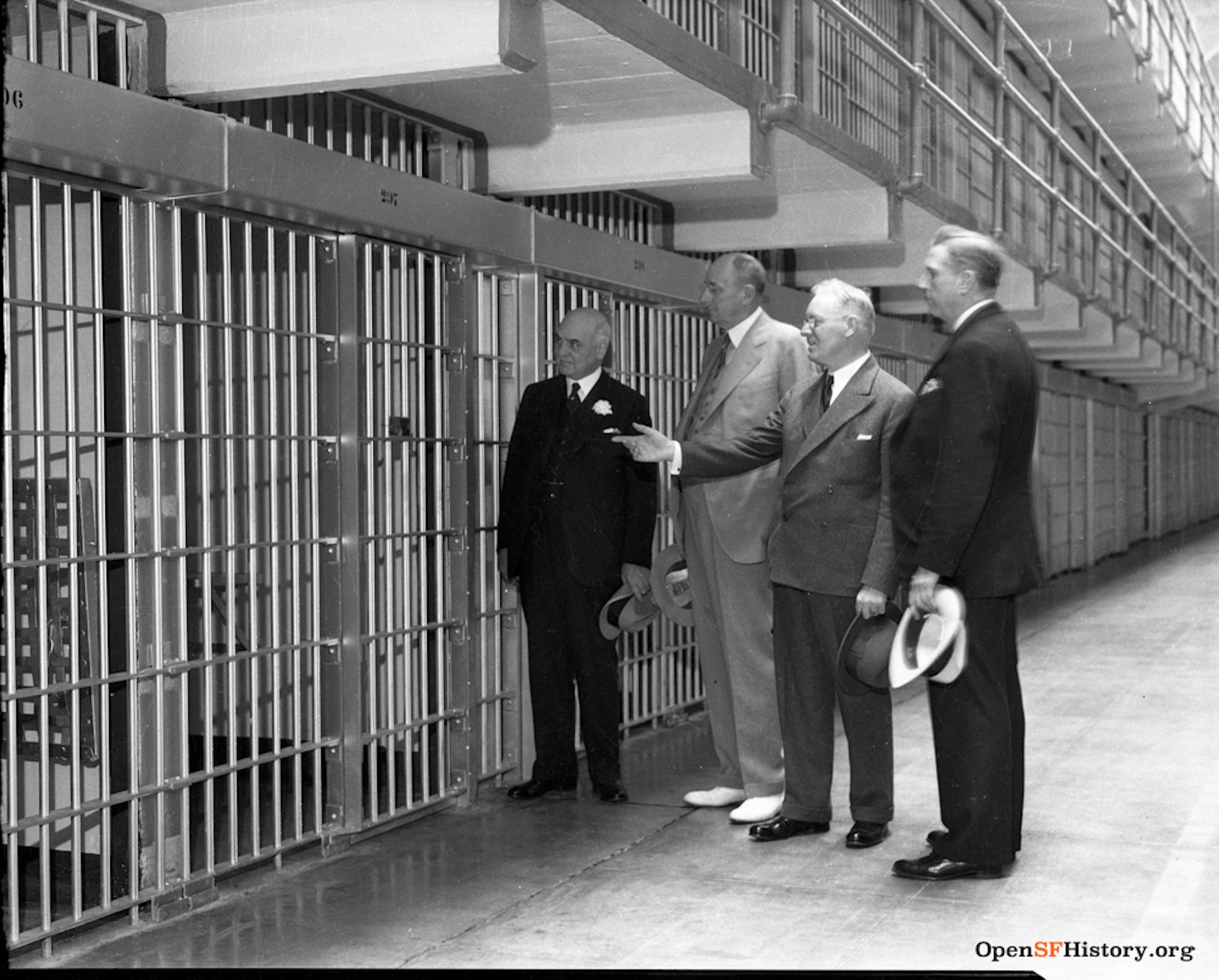 A black and white photograph of four men in 1930s-era suits examining a corridor of prison cells.