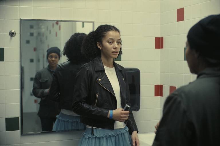 A young Black woman in a white top and black leather jacket faces an older Black woman, whose back is shown to the camera and whose reflection is shown in a bathroom mirror
