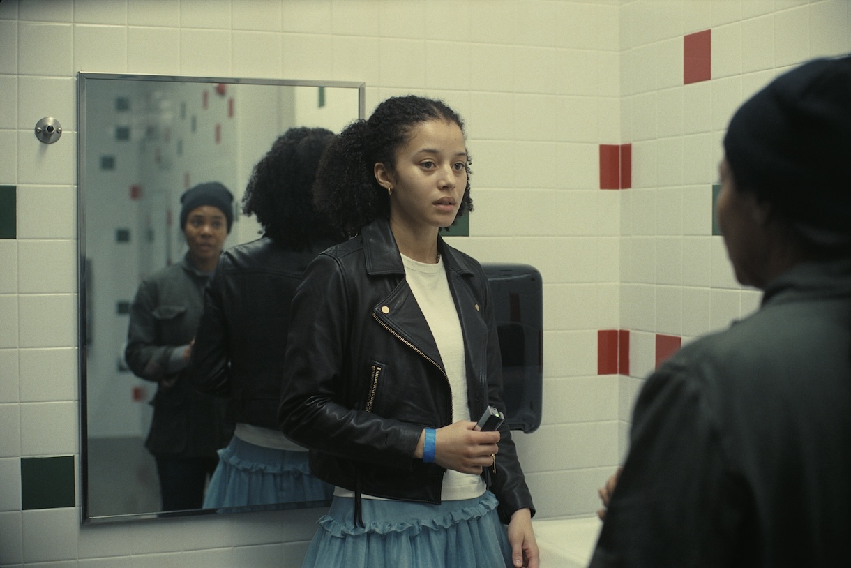 A young Black woman in a white top and black leather jacket faces an older Black woman, whose back is shown to the camera and whose reflection is shown in a bathroom mirror
