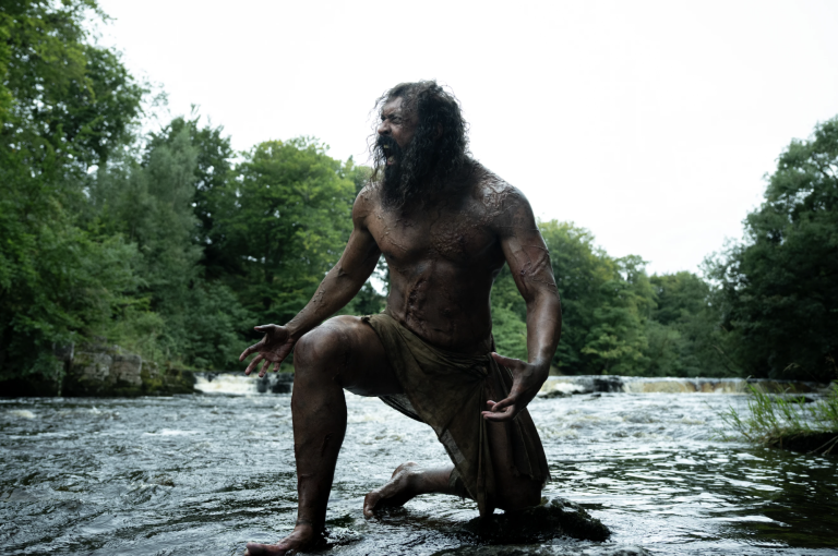 A very muscular man clad only in a loin cloth crouches with one knee on the ground, outside in the wilderness. He is screaming wildly.