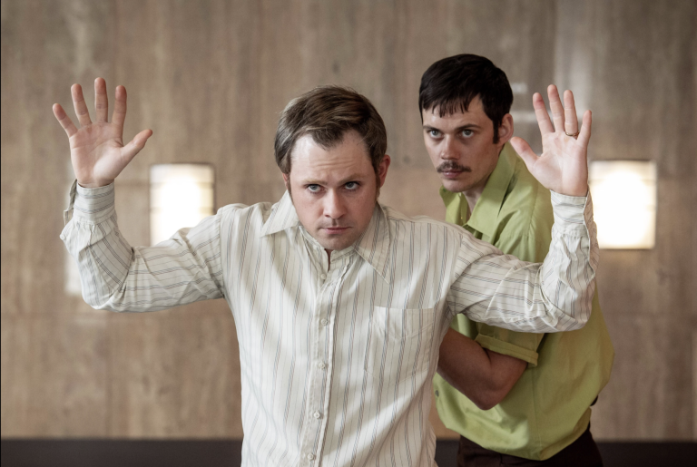 A fearful looking man holds his hands up as another man behind him holds a gun to his back. Both are wearing 1970s-era clothing.