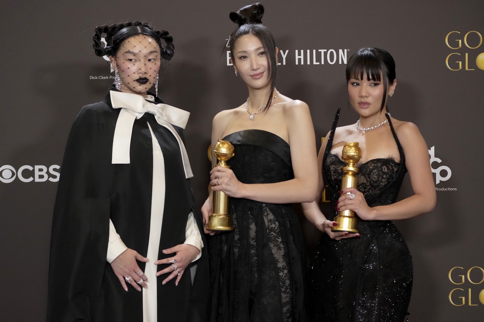 Three glamorous young Asian women stand together on a red carpet, all wearing black evening gowns. Two are holding Golden Globe awards.