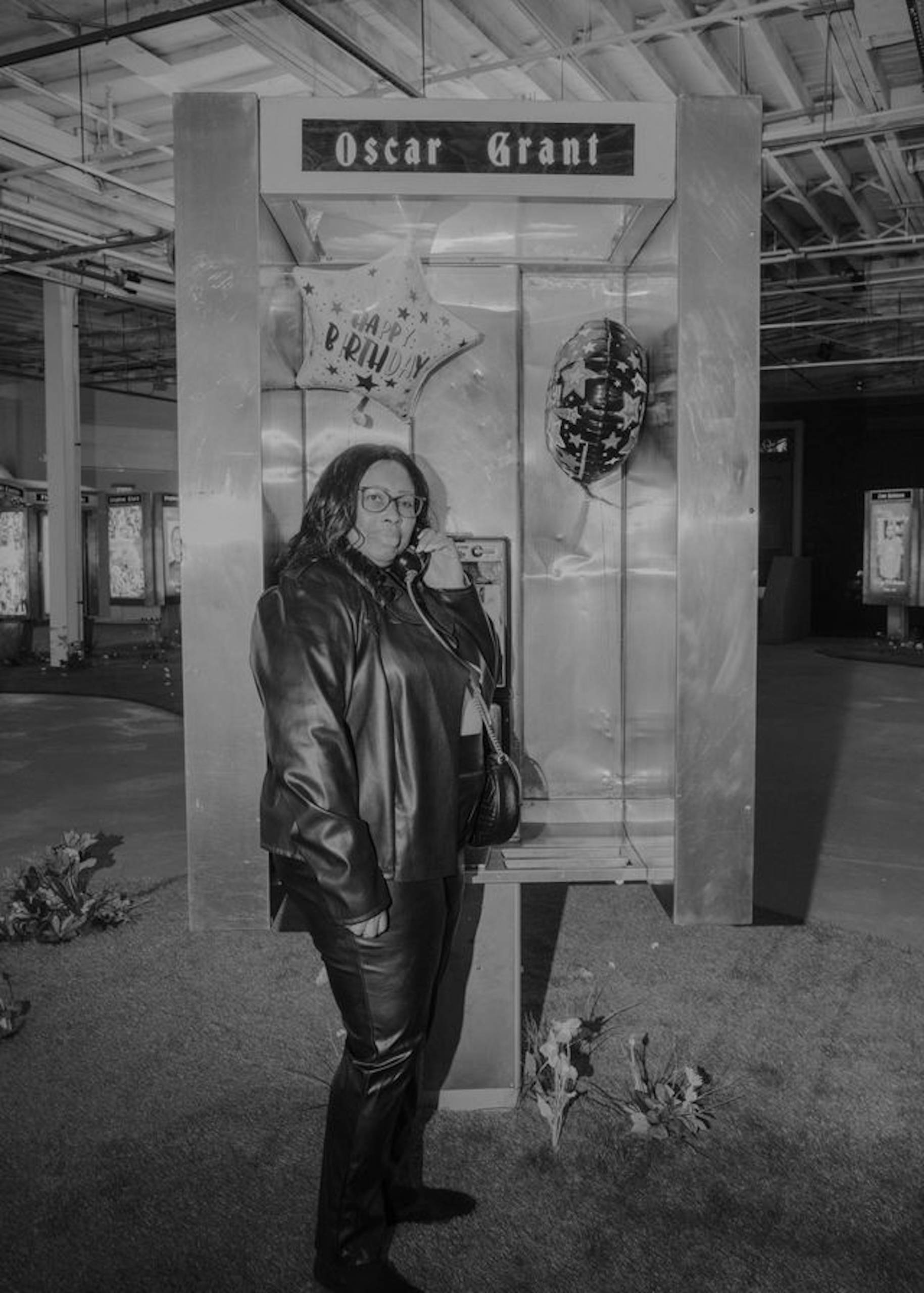 A black and white image of an African American woman in a black leather jacket and glasses standing at a phone booth where two "happy birthday" balloons are mounted. 