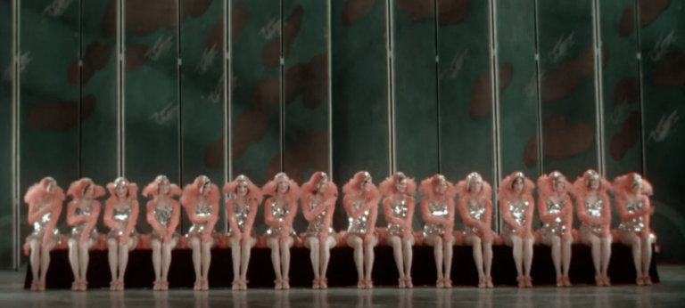 A film still showing a seated line of 16 dancers, all wearing pink curly wigs and shimmering silver costumes.