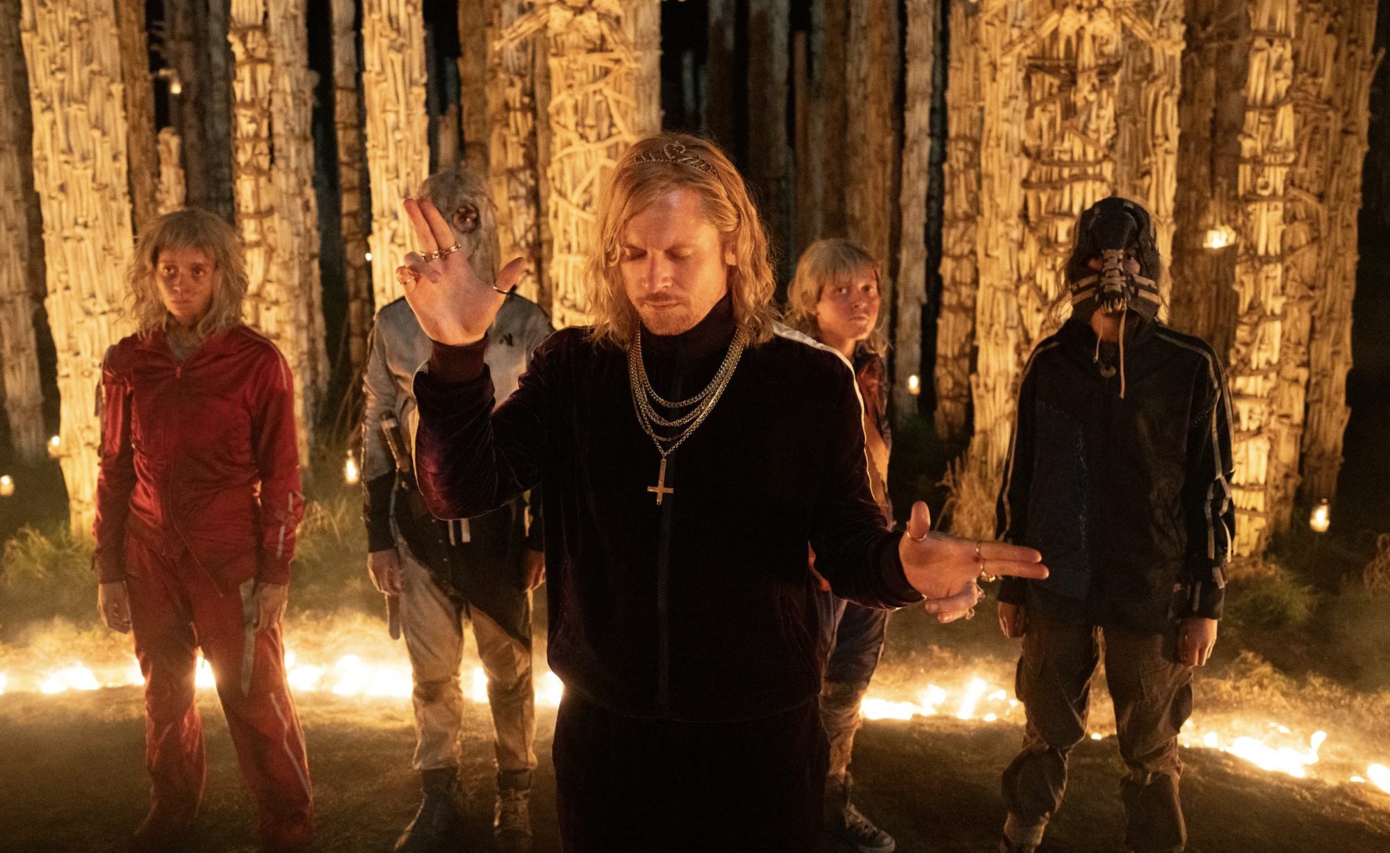 A blond man wearing a black tracksuit stands, eyes closed, hands gesturing, in a strange spotlit circle. Behind him are four young people, also wearing tracksuits, and looking the worse for wear.