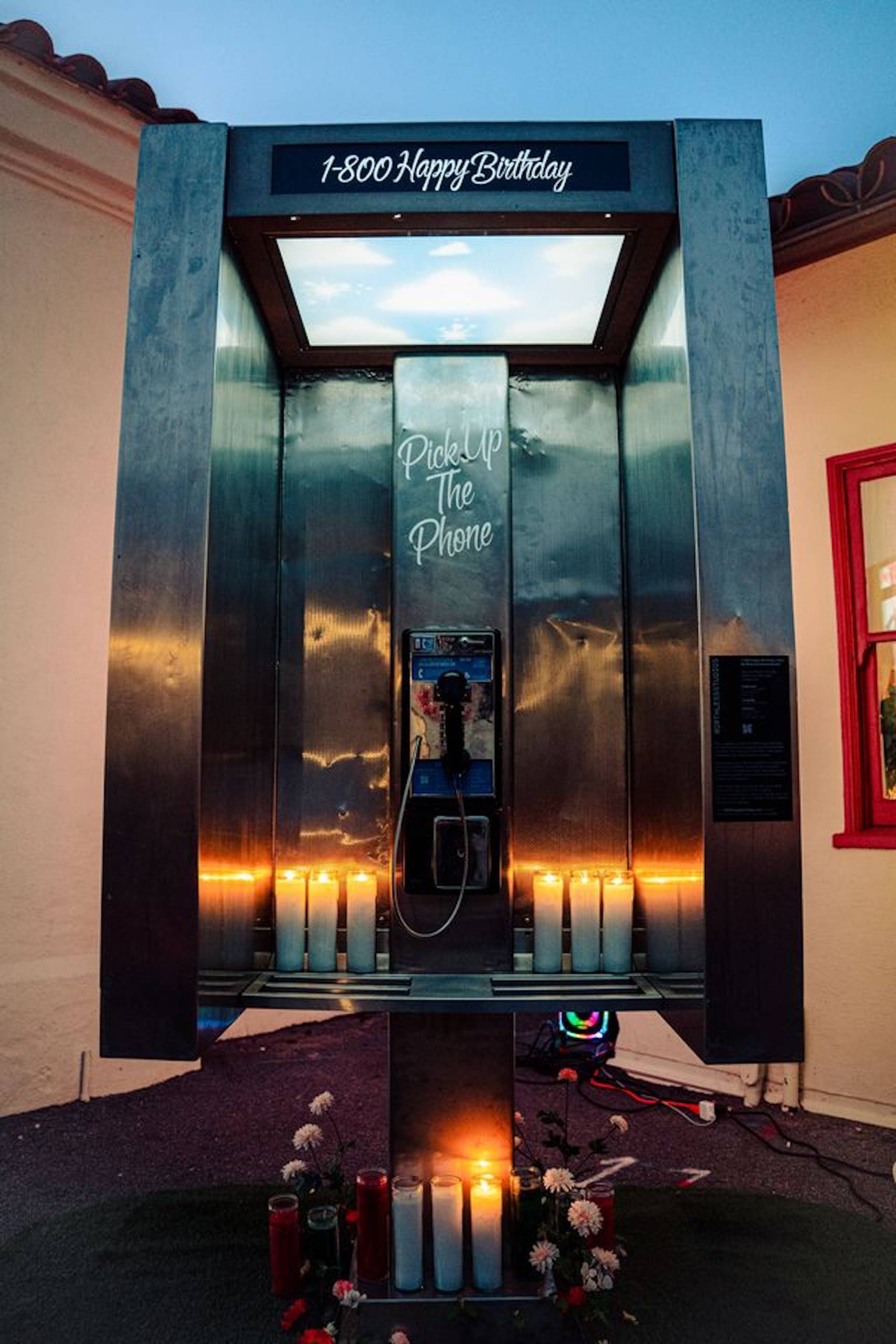 A photo of a payphone booth surrounded by lit candles, the words "1-800 Happy Birthday" inscribed at the top.