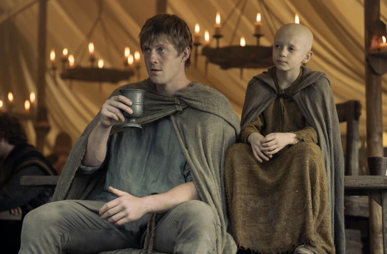 A white man and young boy, both dressed in medieval clothing, sit side by side in a tent lit by candles.