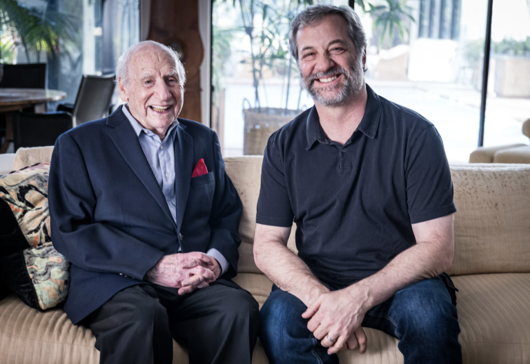 An elderly man wearing a suit with no tie sits on a beige couch smiling broadly. At his side is a middle aged man in jeans and a polo shirt, also laughing.