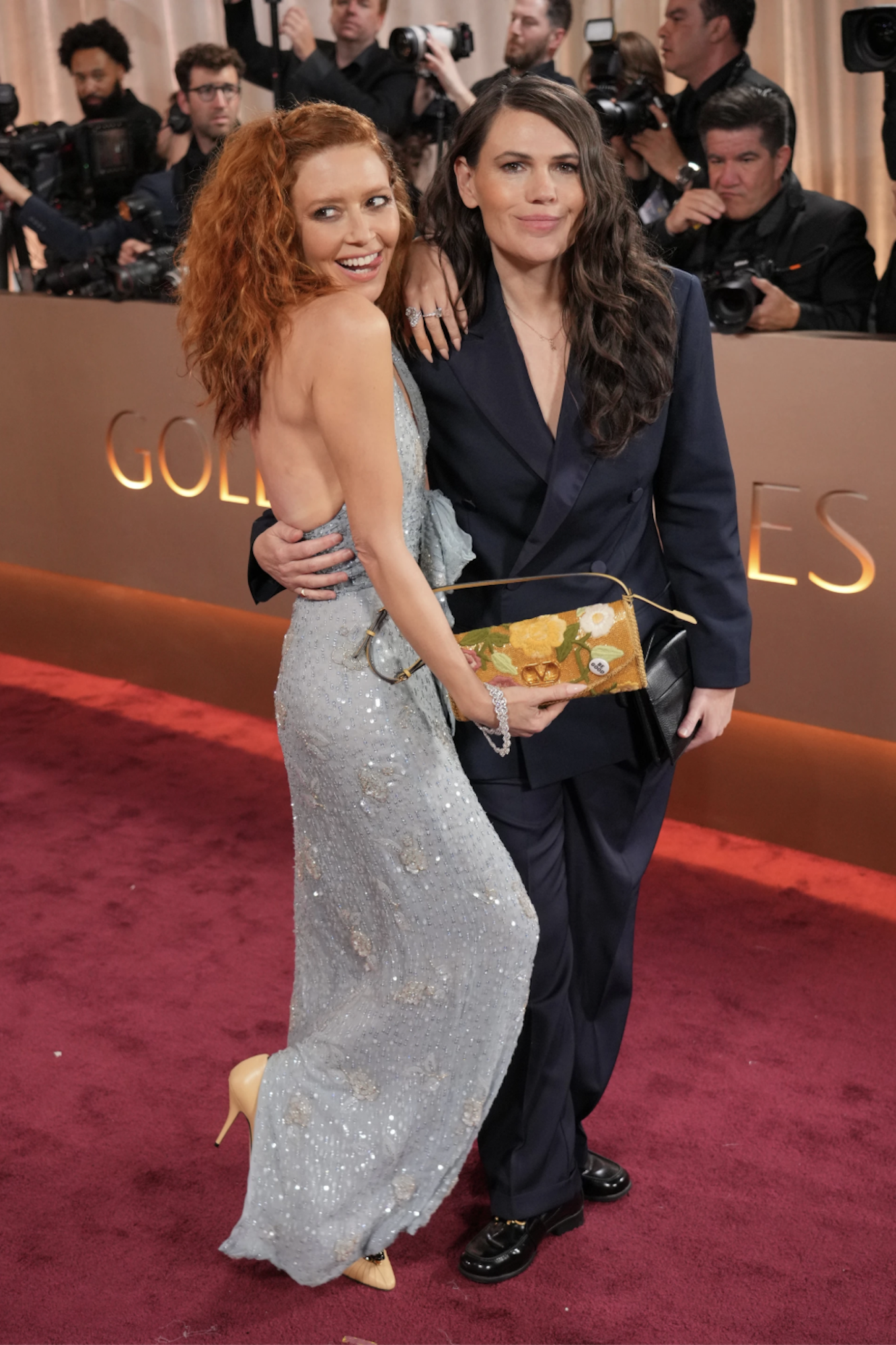A white woman with dark hair in a navy blue suit stands with her arm around the waist of a vivacious white woman in a backless pale blue gown, holding a clutch purse with a pin on it.