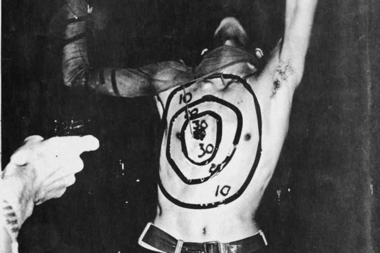 A book cover featuring a black and white photograph of a thin, white man, performing on his knees, his face obscured, with a target drawn on his shirtless torso. An audience member's arm reaches out with their hand positioned as if pulling a trigger.