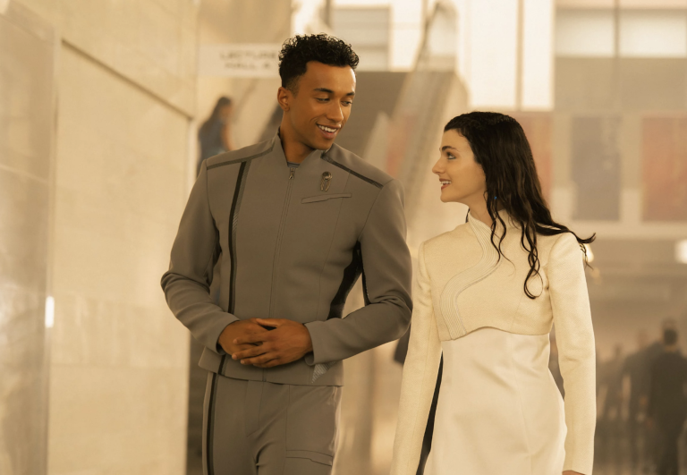 A young Black man in a grey uniform walks through white corridors alongside a young white woman with long black hair in a white uniform. They are smiling at one another.