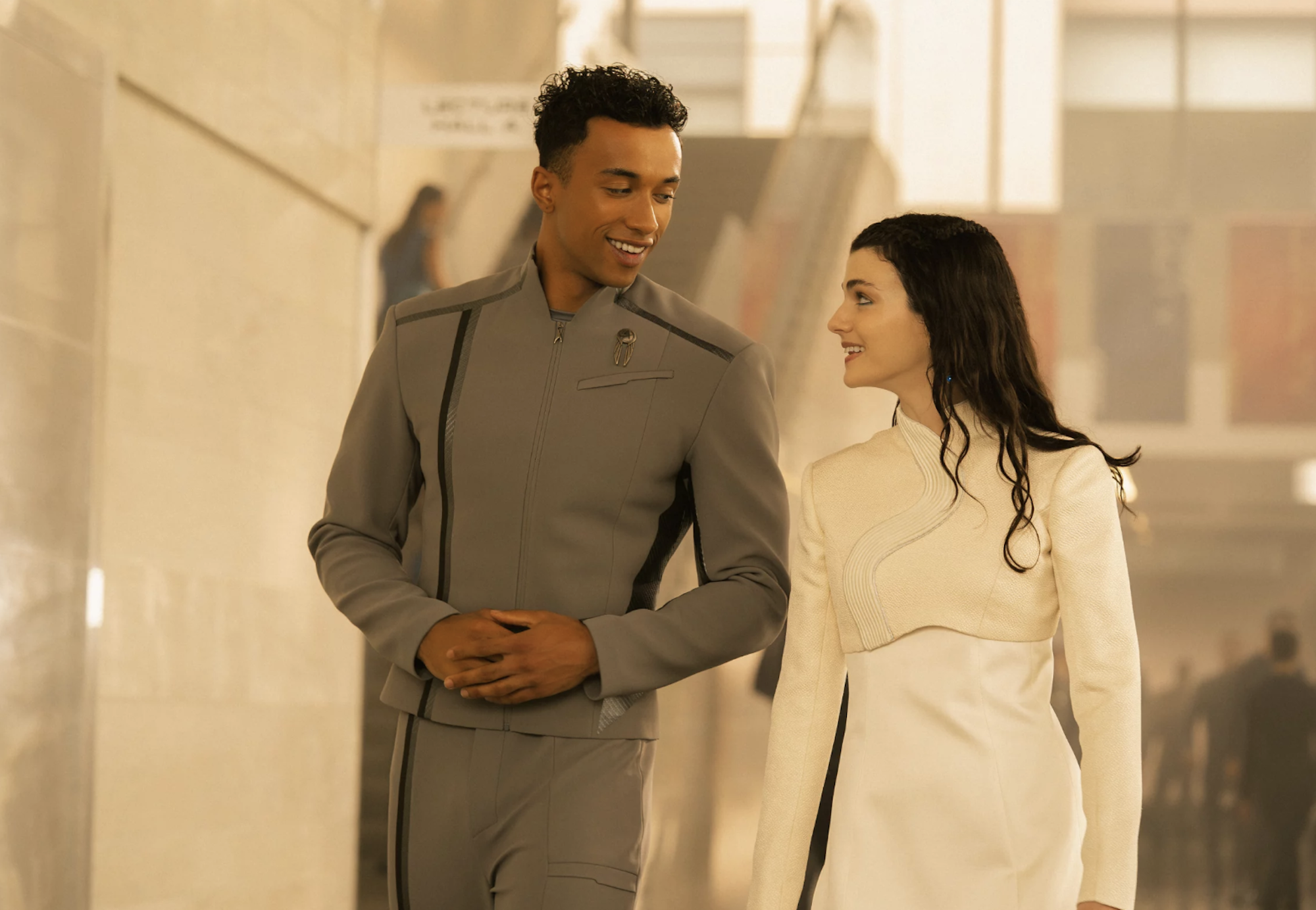 A young Black man in a grey uniform walks through white corridors alongside a young white woman with long black hair in a white uniform. They are smiling at one another.