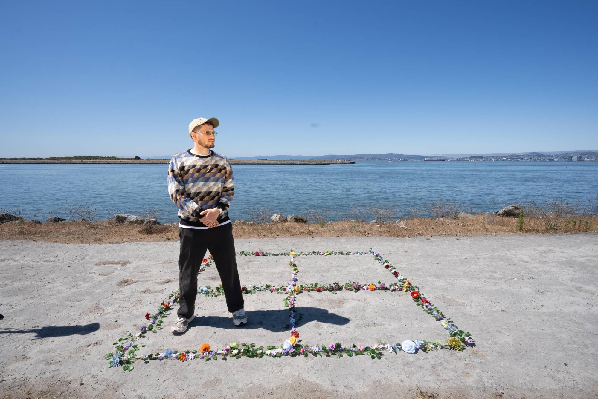 A man in a hat and colorful sweater poses for a photo while standing near the San Francisco Bay, surrounded by flowers in the shape of four squares.