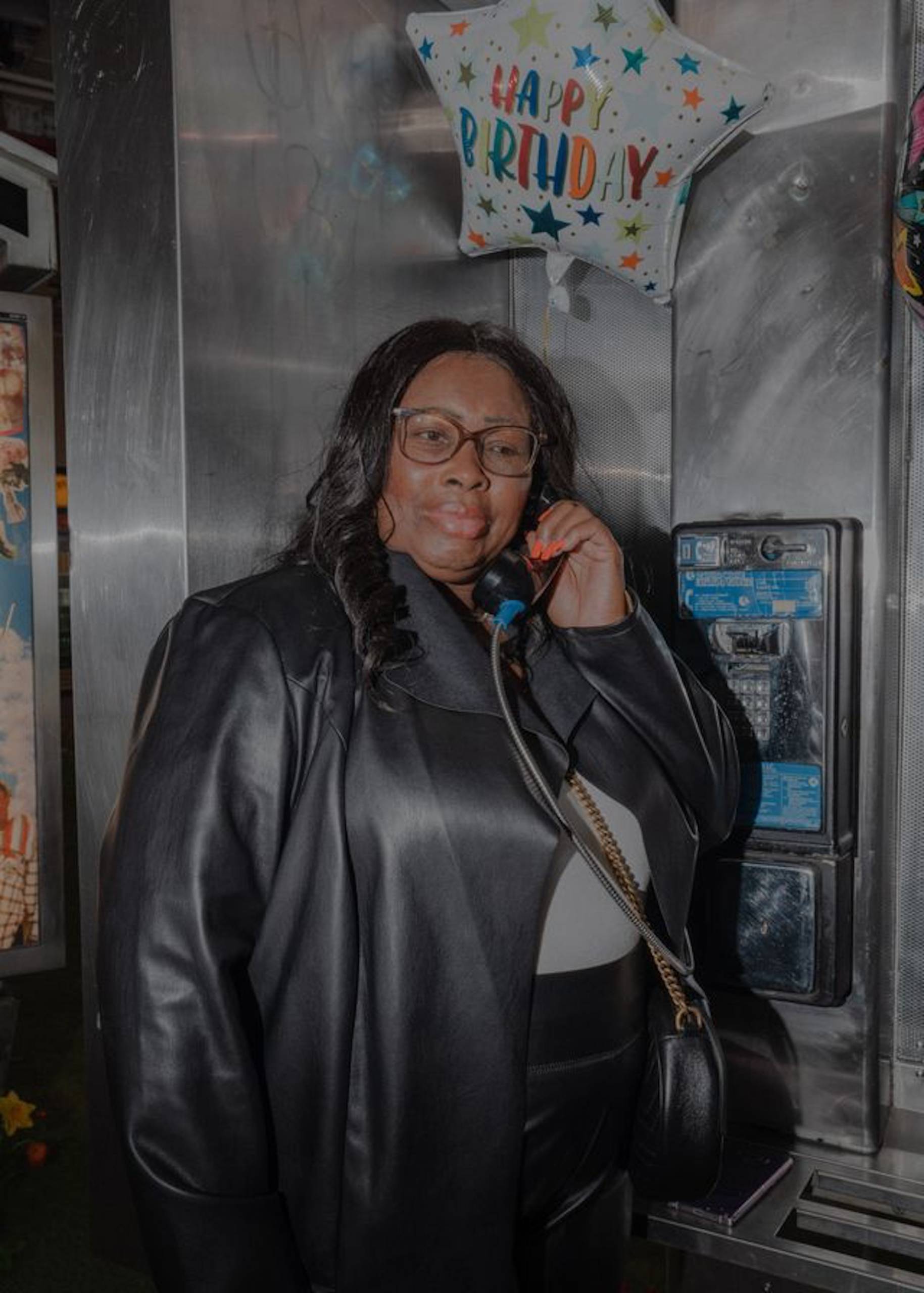 An African American woman wearing glasses and a leather jacket stands at a phone booth holding a phone and looking into the distance.