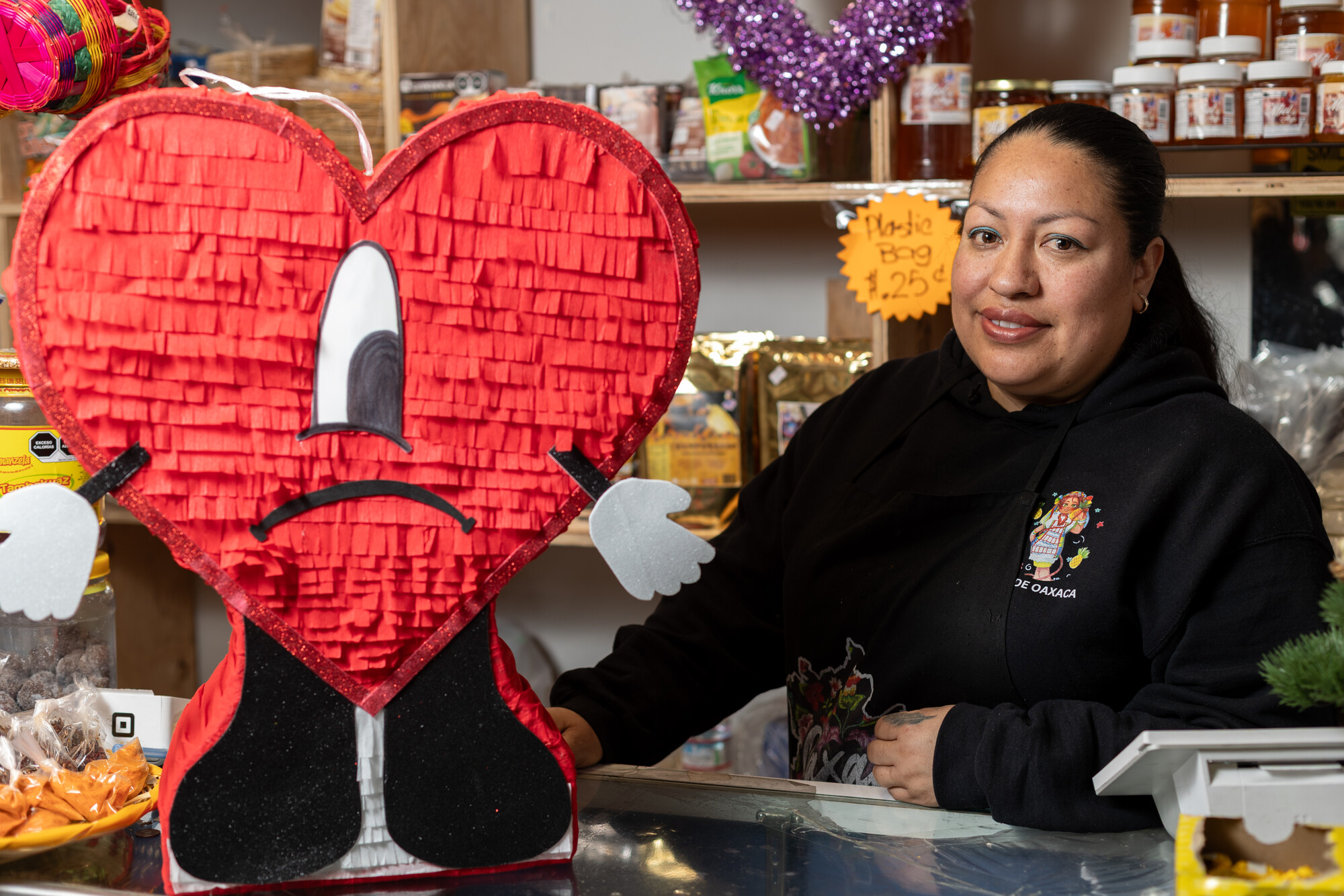 A Latina woman in black top poses next to a large red heart-shaped pi&ntilde;ata, with shelves of merchandise in the background