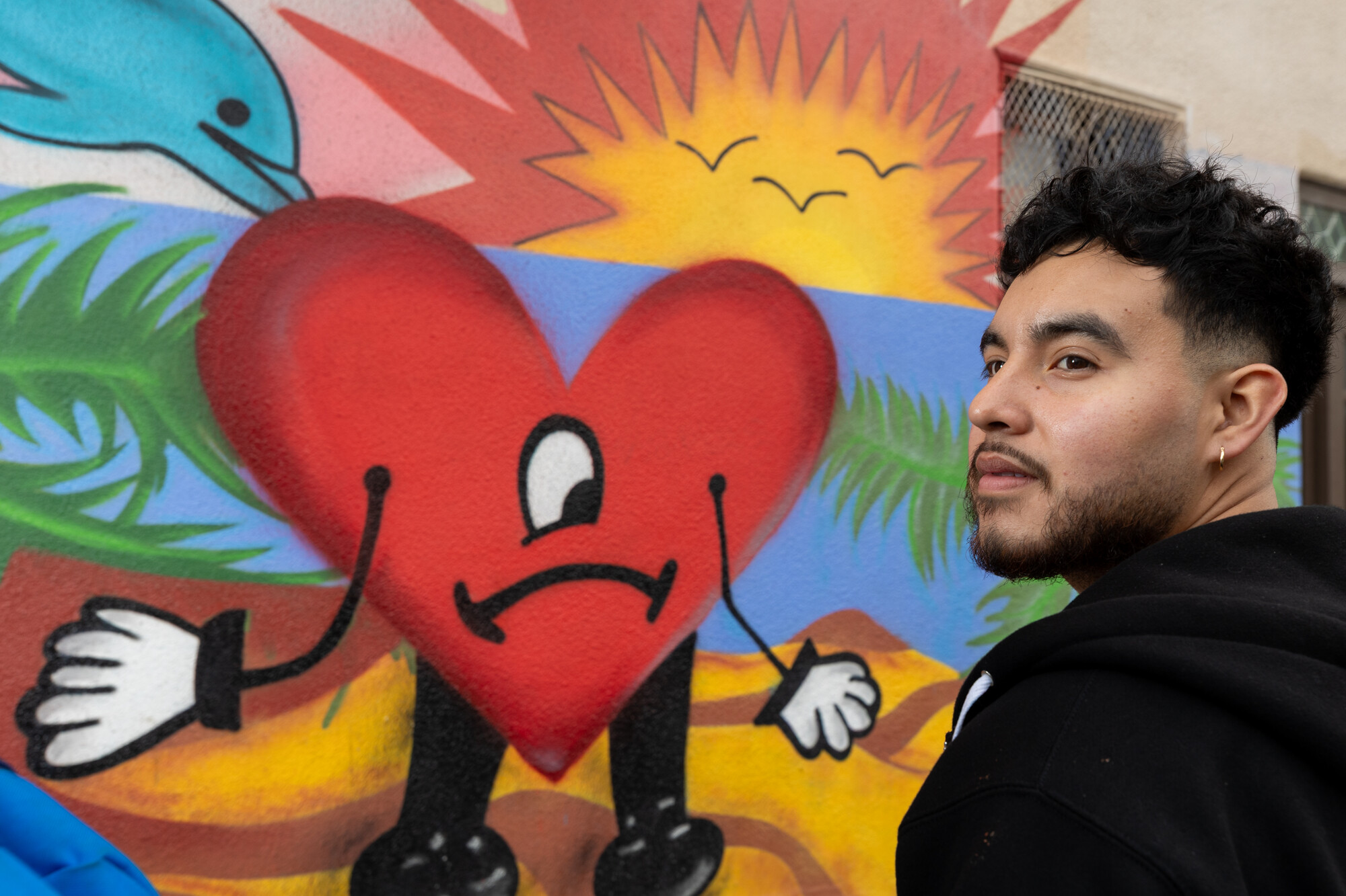 A Latino man with short black hair and a beard, in a black top, stands in front of a colorful mural of a heart with a sad face, a sunset, and dolphins