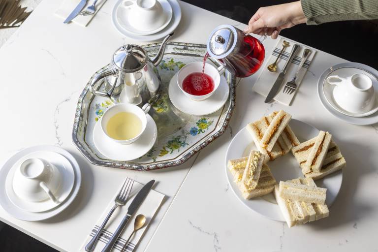 A woman's hand is seen pouring red hibiscus tea into a porcelain tea cup. On the table is a plate of finger sandwiches.