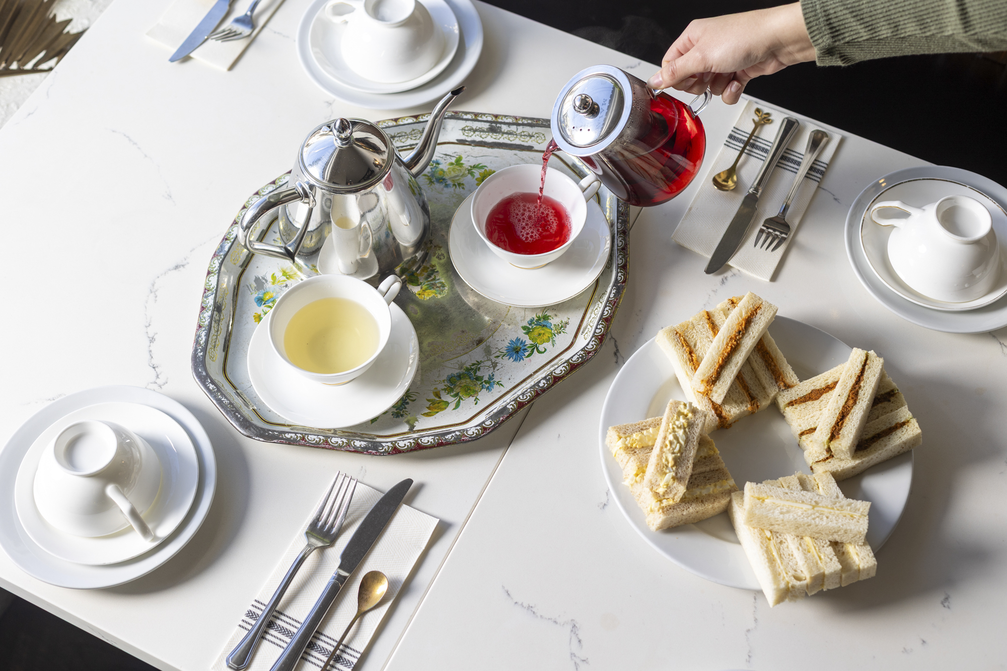 A woman's hand is seen pouring red hibiscus tea into a porcelain tea cup. On the table is a plate of finger sandwiches.