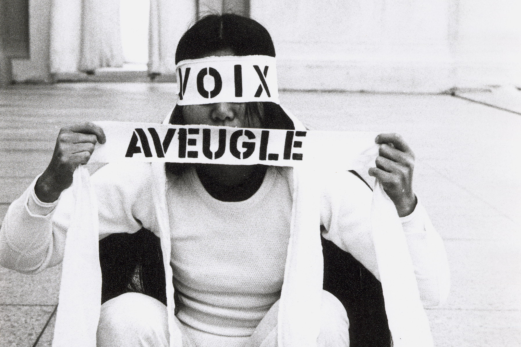 black and white image of woman squatting in white with eyes and mouth covered by 'voix' and 'aveugle'
