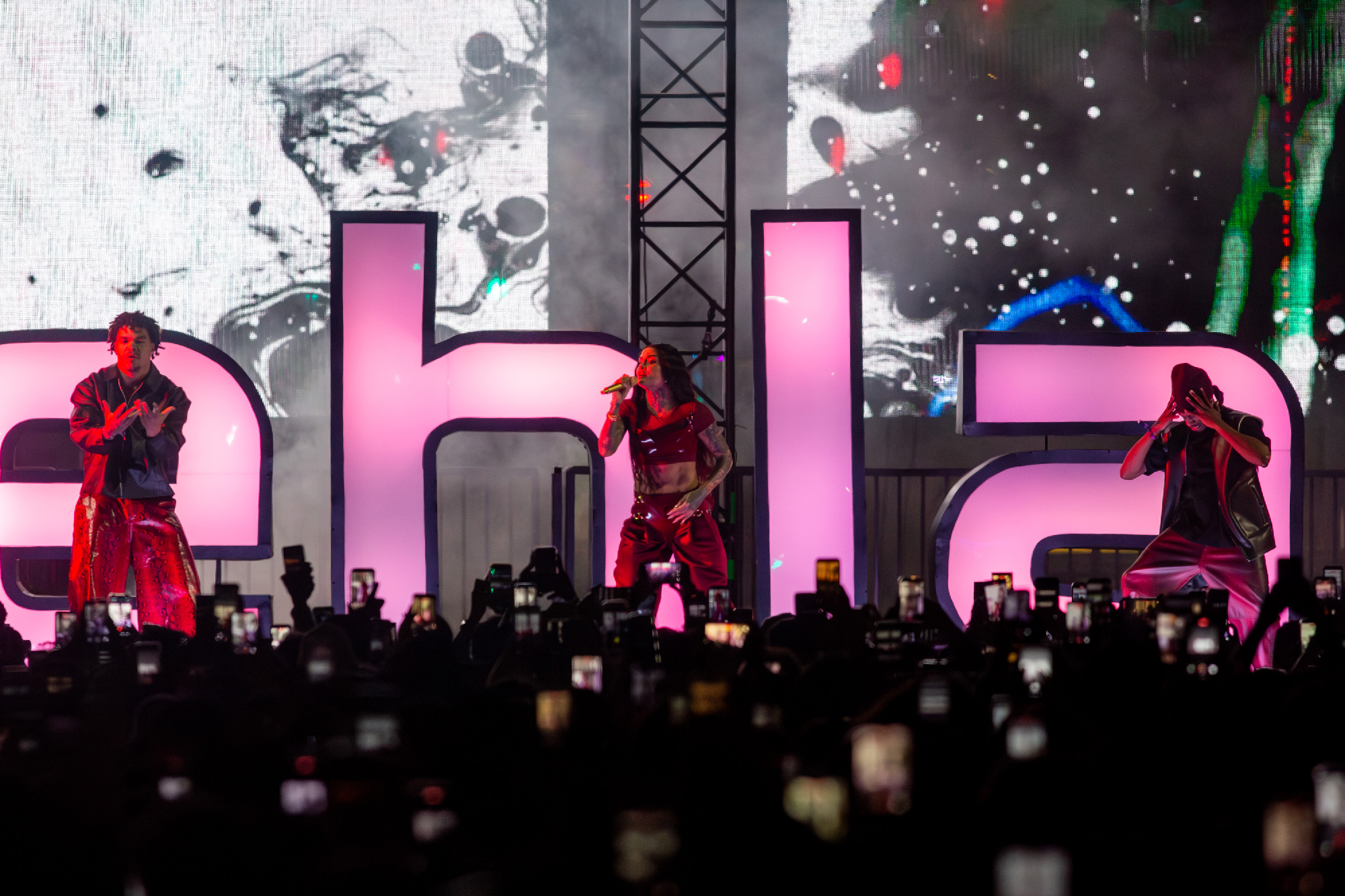 singer and two dancers on stage with crowd holding phones
