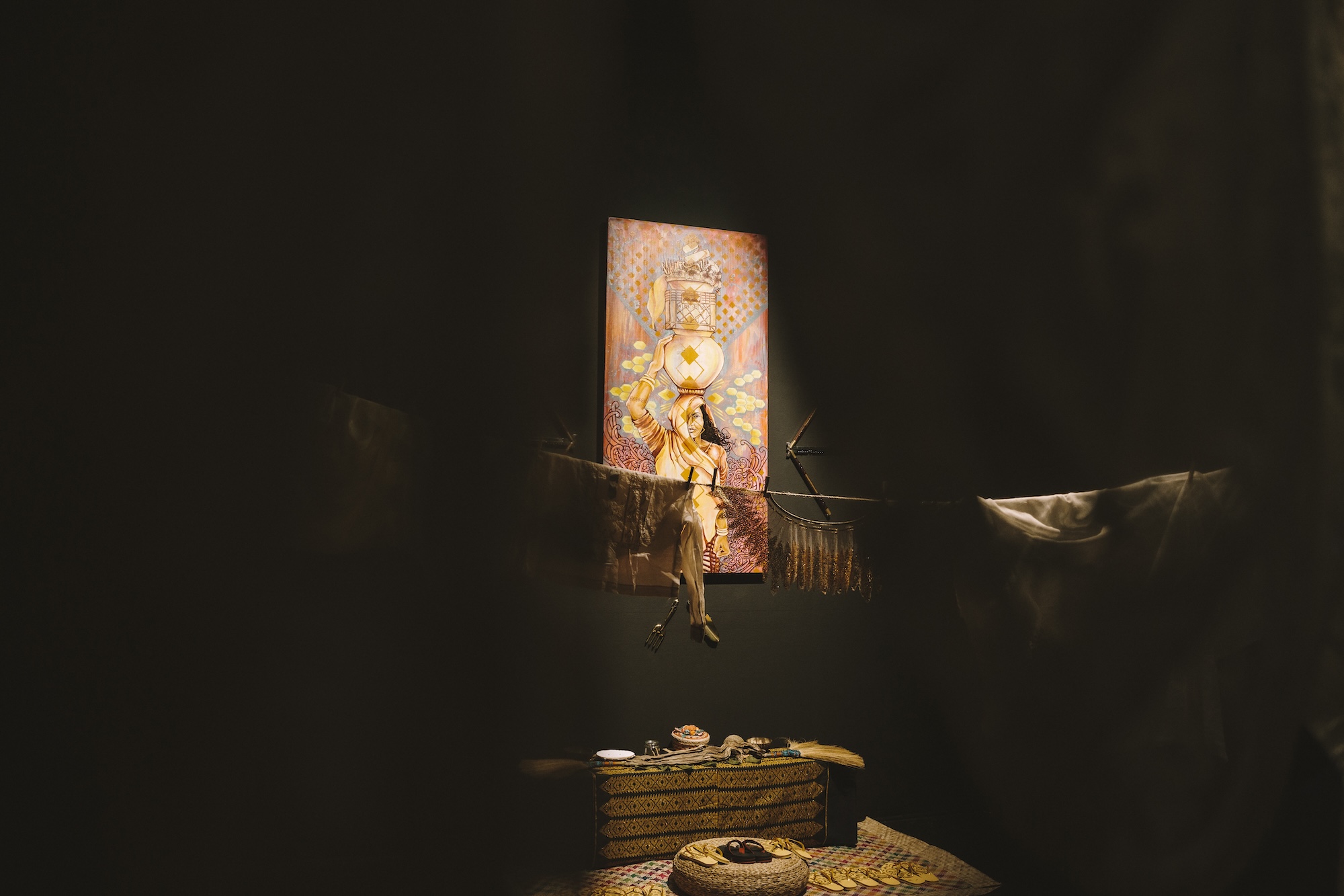 Through white clothing on a clothes line a painted picture is revealed, depicting a woman balancing a gourd on her head with one hand. 