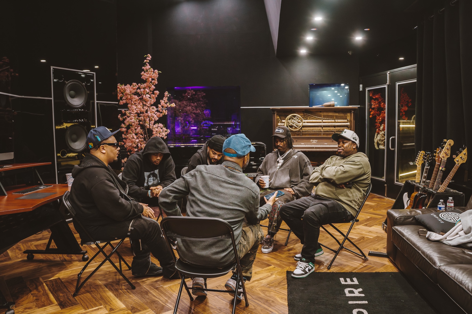 Six African American men sit in a circle while holding a conversation in a music studio 