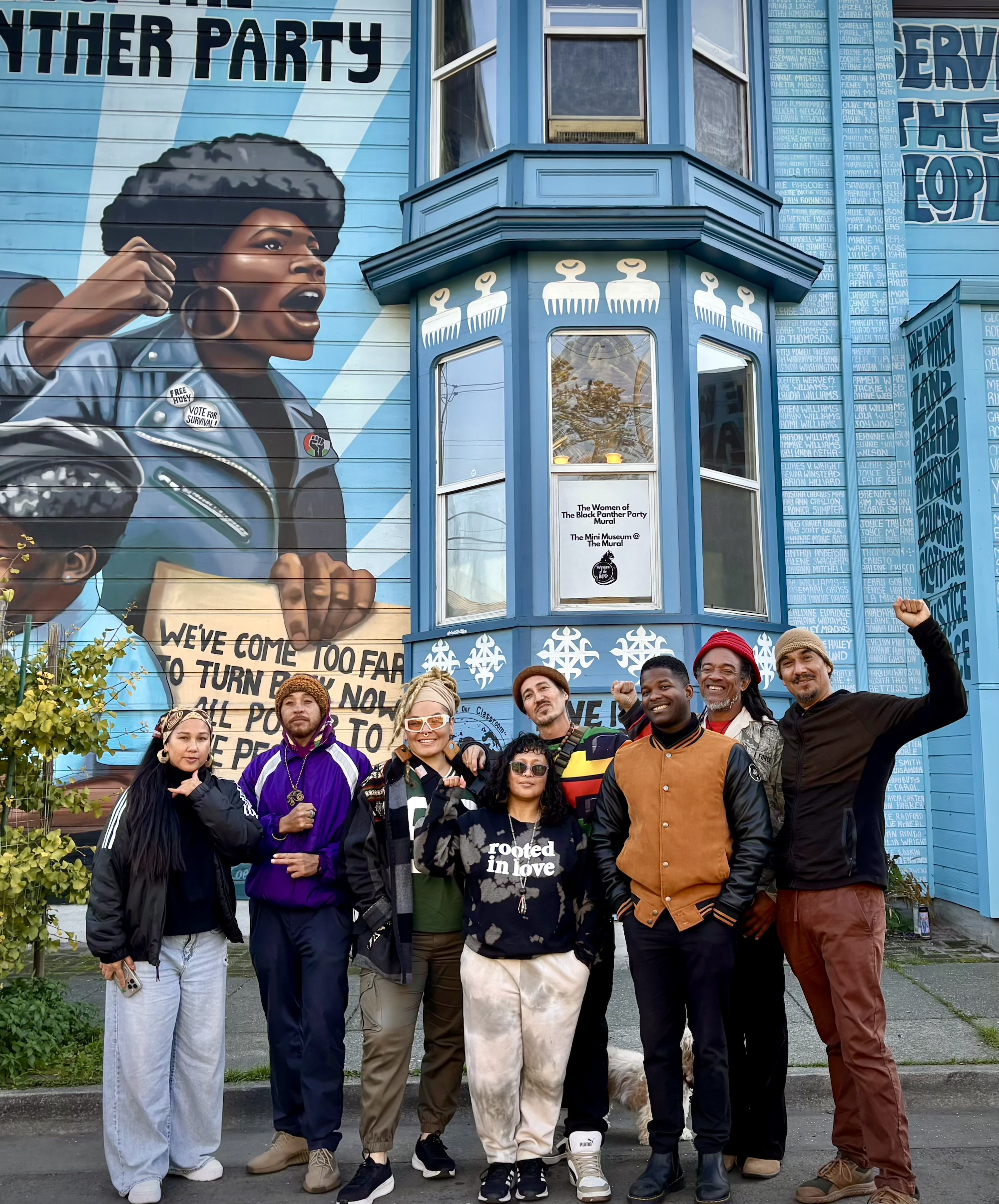 Eight people posing for a photo outside, in front of a building with a mural painted on it.