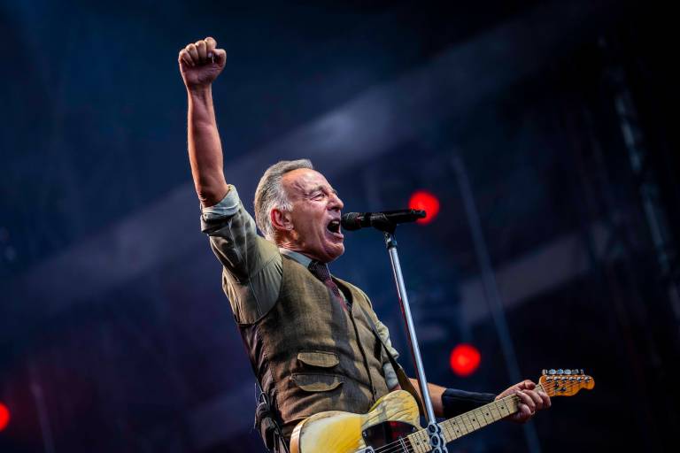 A man in a collared shirt and vest holds a Fender telecaster guitar and sings into a microphone with his fist in the air