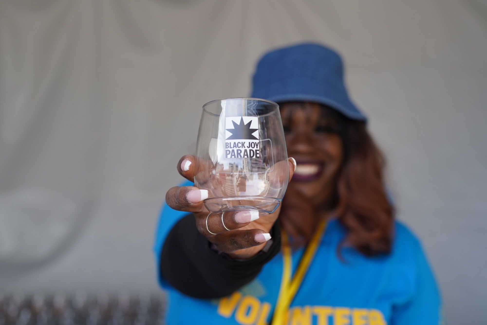 Woman in blue shirt and blue hat holds up a wine glass that reads "Black Joy Parade"