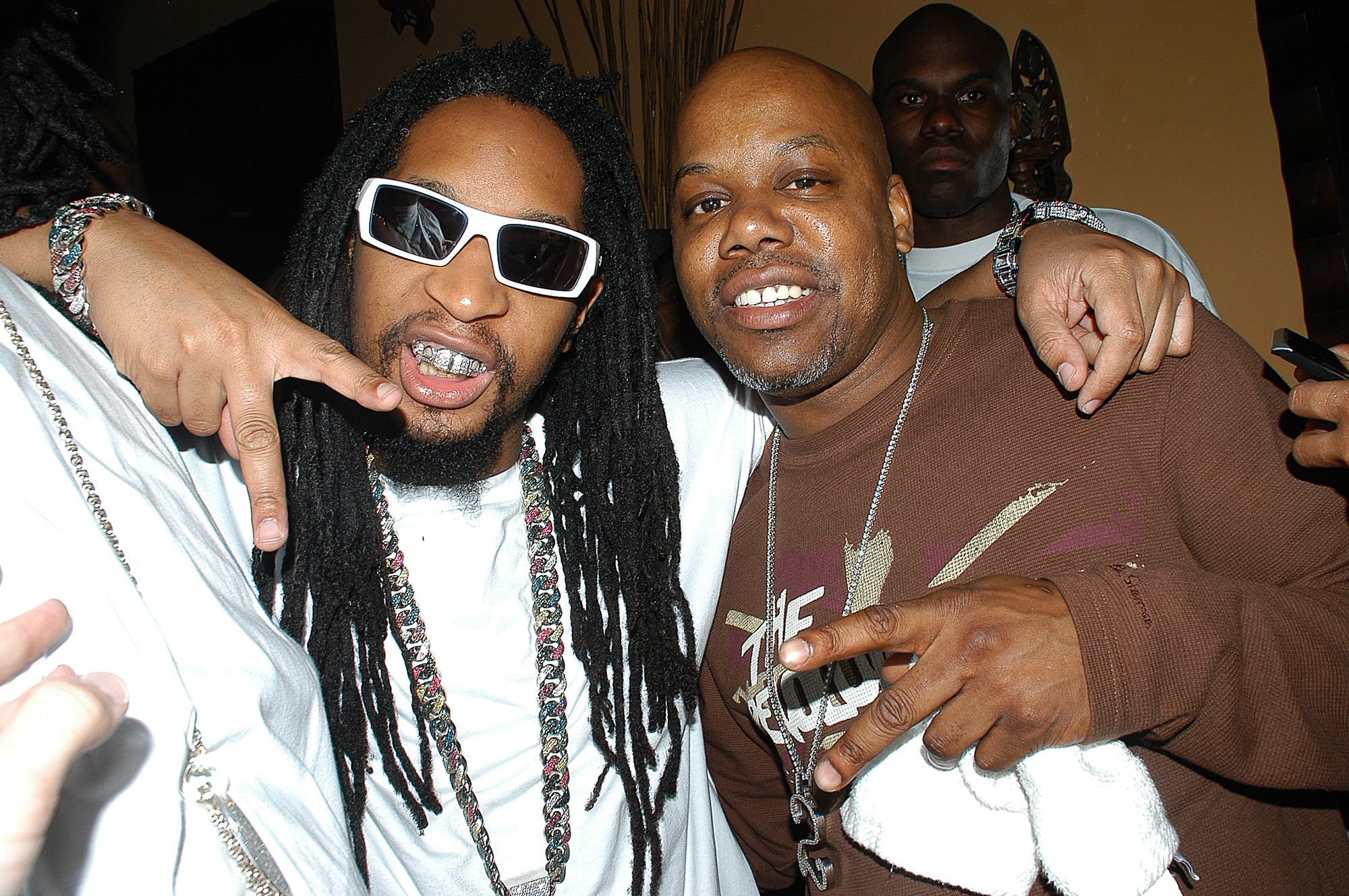 Two Black men, one in white with dreadlocks and one in brown and bald, embrace and smile for the camera