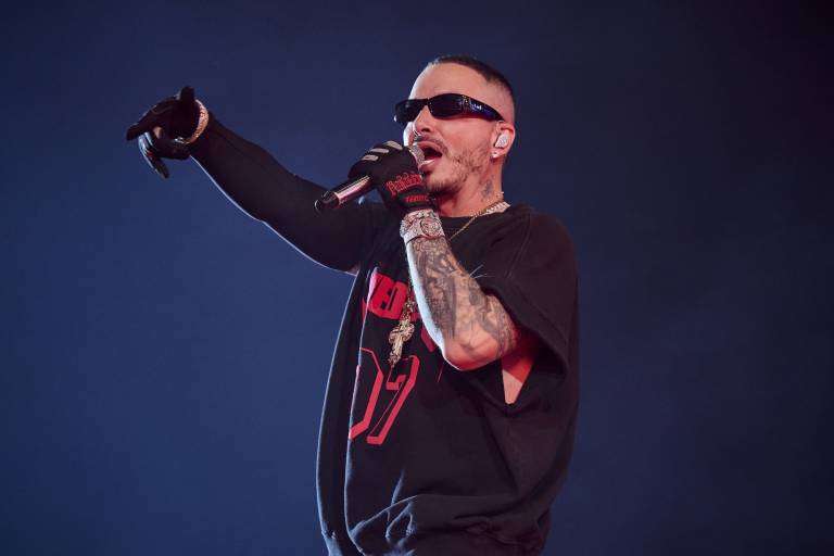 A man in wrap-around sunglasses, black gloves and a black and red T-shirt sings into a microphone against a blue background