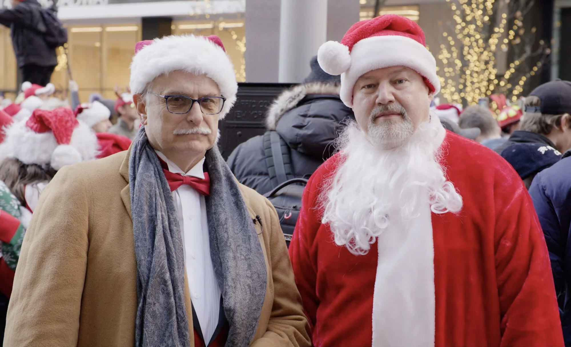 A distinguished older white man with a white mustache wearing a red bow tie, santa hat and camel colored winter coat, stands next to a white man of similar age wearing a santa suit.