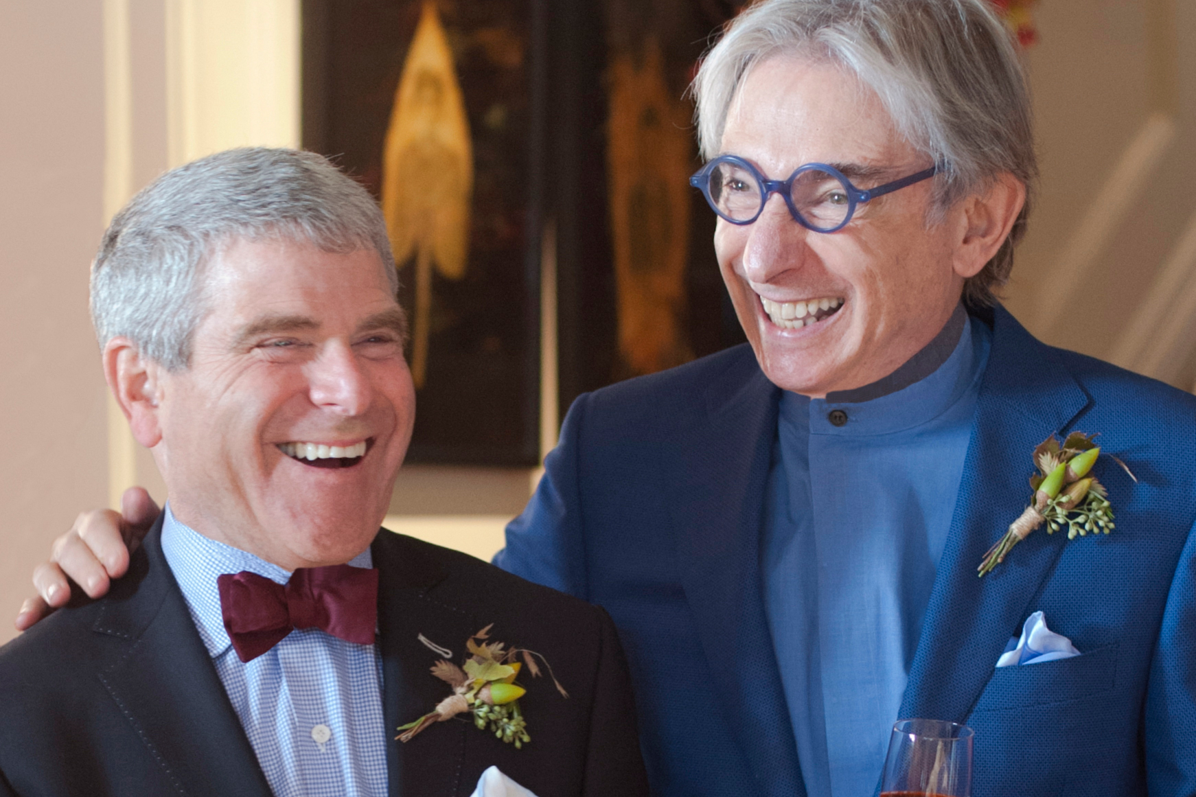 A man in a black suit and deep maroon bowtie smiles next to a man in blue suit and glasses