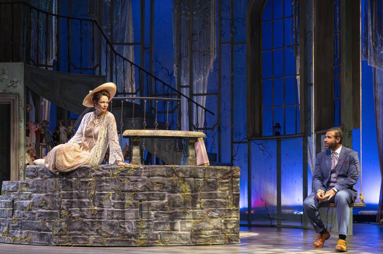 A white woman in a dress and large hat sits on a stone wall, looking to the right at a Black man in a blue suit, sitting on a swing